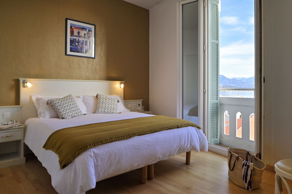 A neatly made bed with a white duvet, a mustard-colored throw, and patterned pillows. A framed picture hangs above the bed.