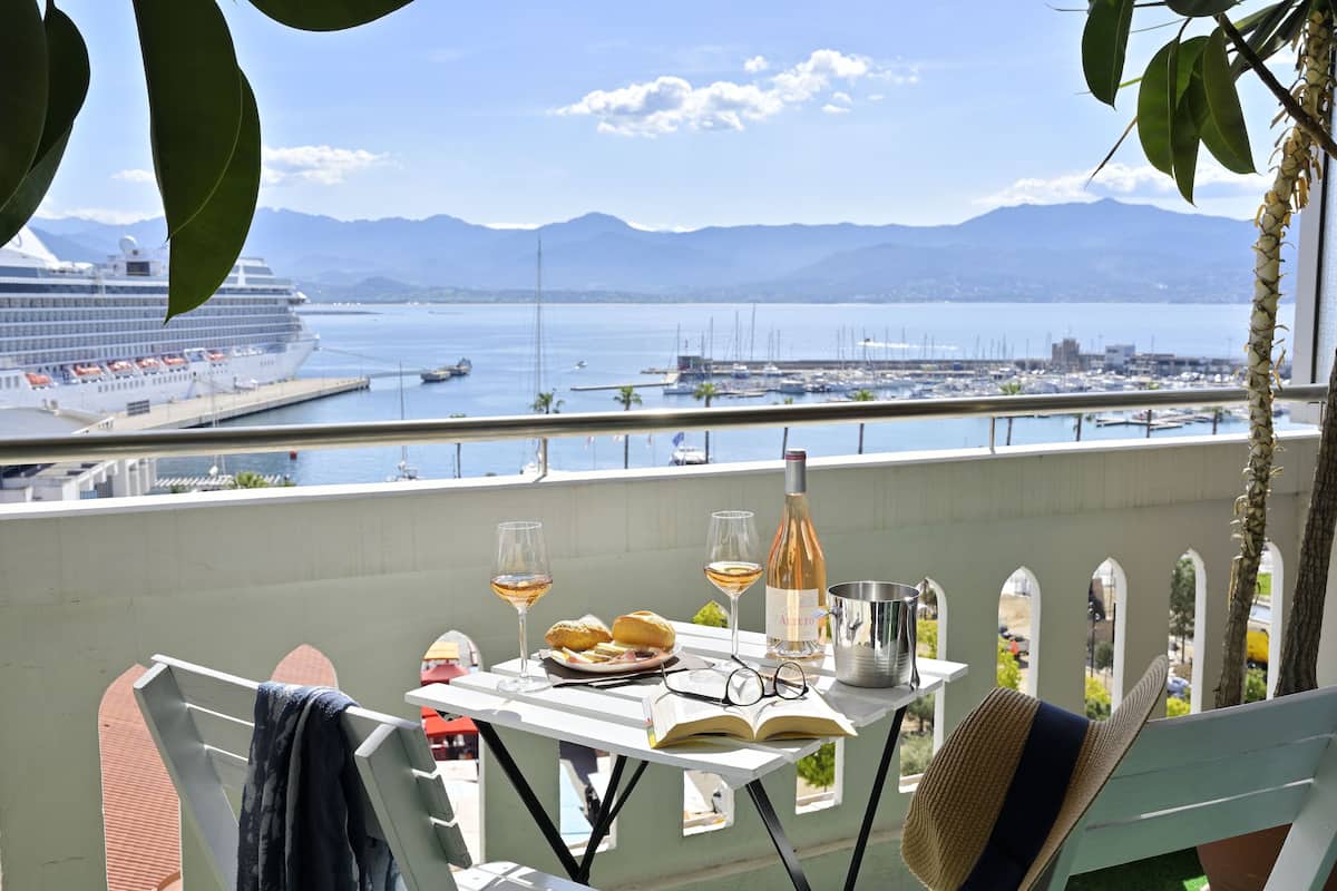 A balcony with a table set for two, featuring a bottle of wine, glasses, a plate of food, and a book.