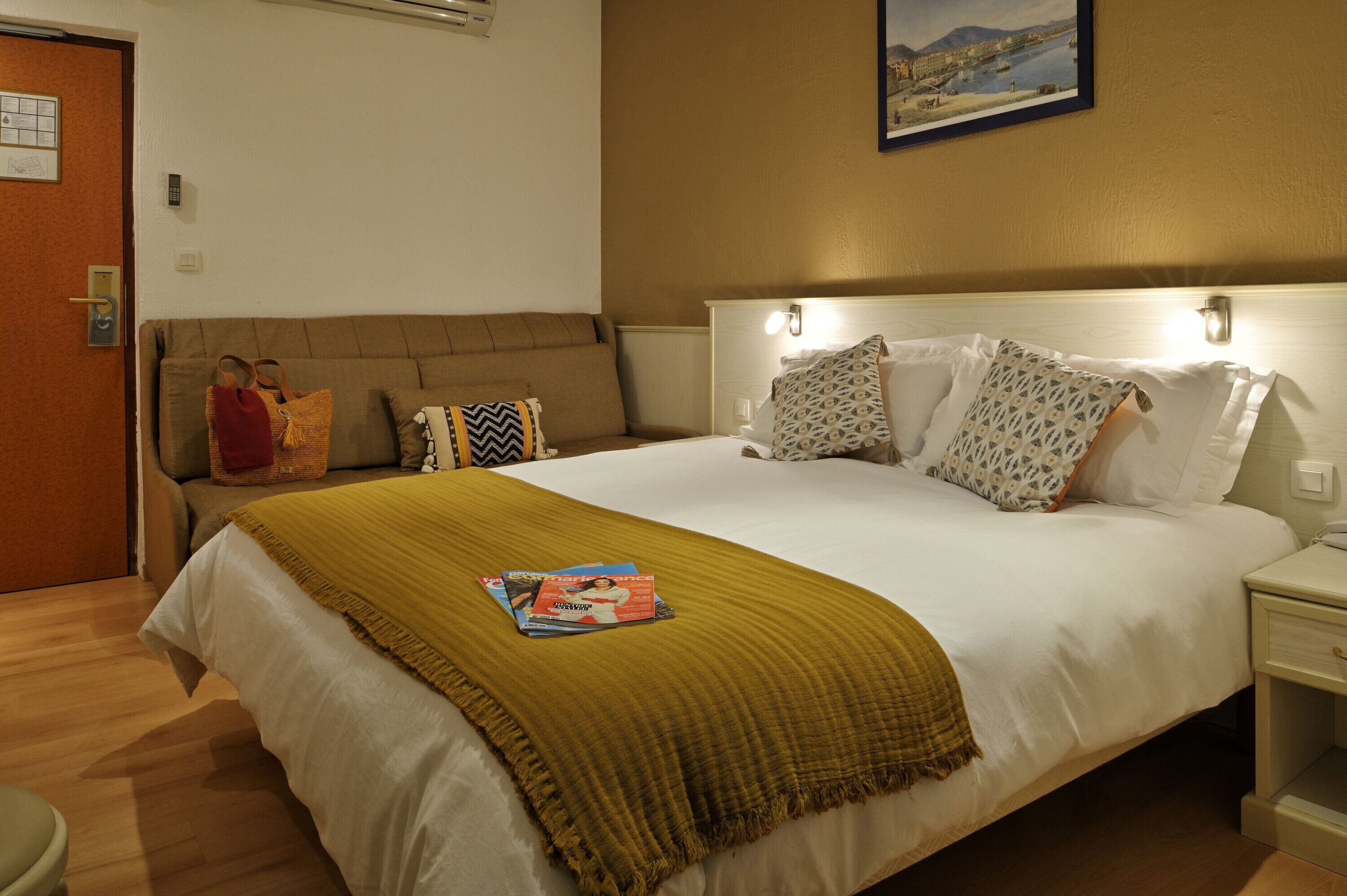 A hotel room with a bed, a nightstand, a wall-mounted lamp, a painting, and a door with a remote control.