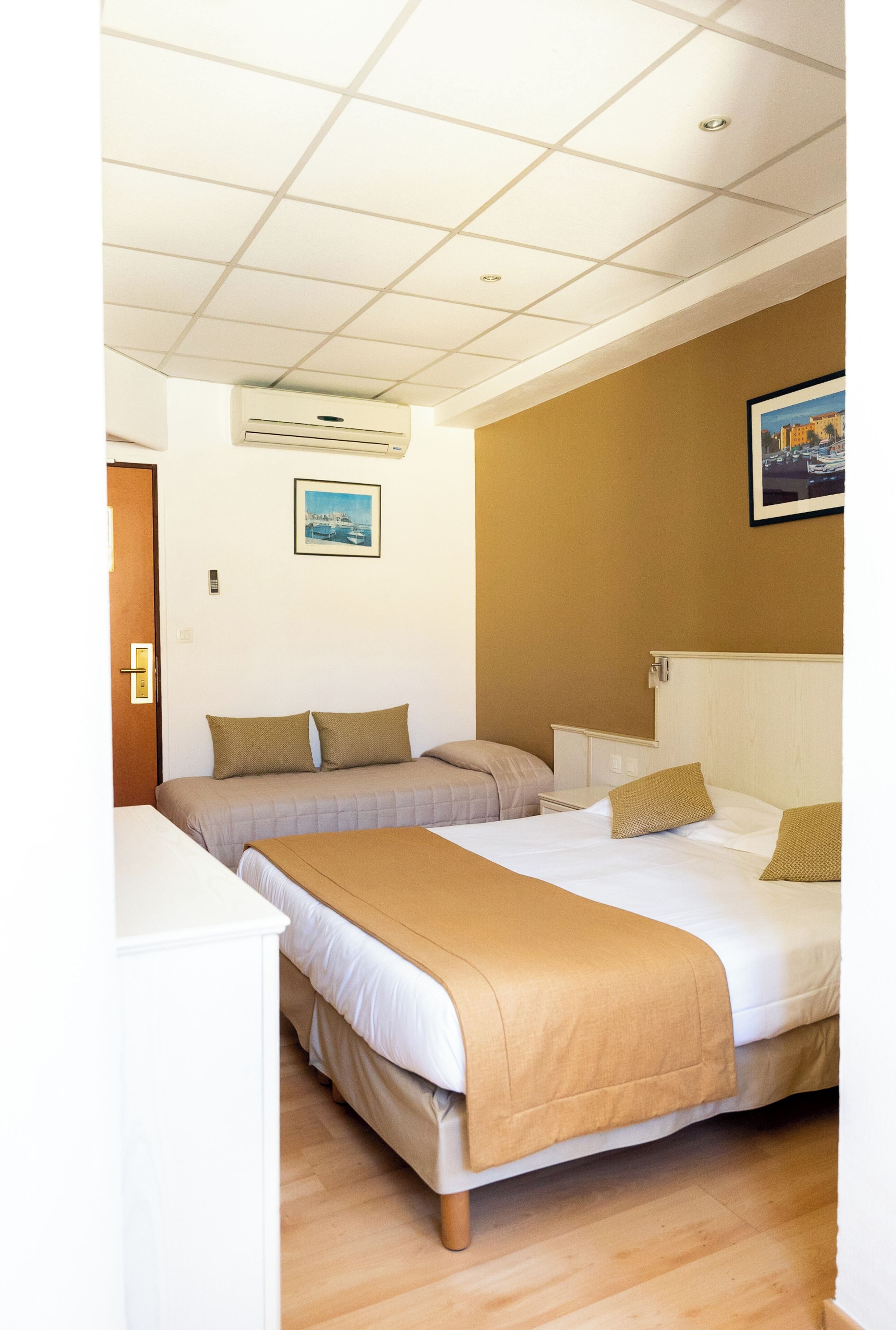 A hotel room with two beds, a white wardrobe, a painting on the wall, and an air conditioning unit.
