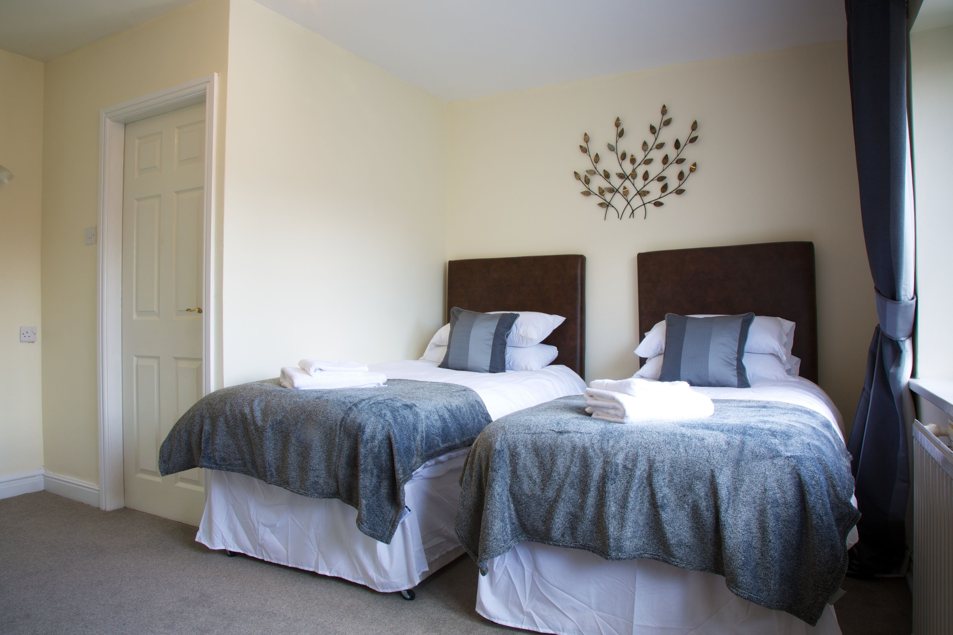 A hotel room with two beds, each with a grey blanket and white sheets. There is a decorative wall piece above the beds and a white door on the left.