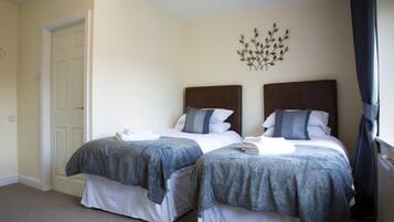 A hotel room with two beds, each with a grey blanket and white sheets. There is a decorative wall piece above the beds and a white door on the left.