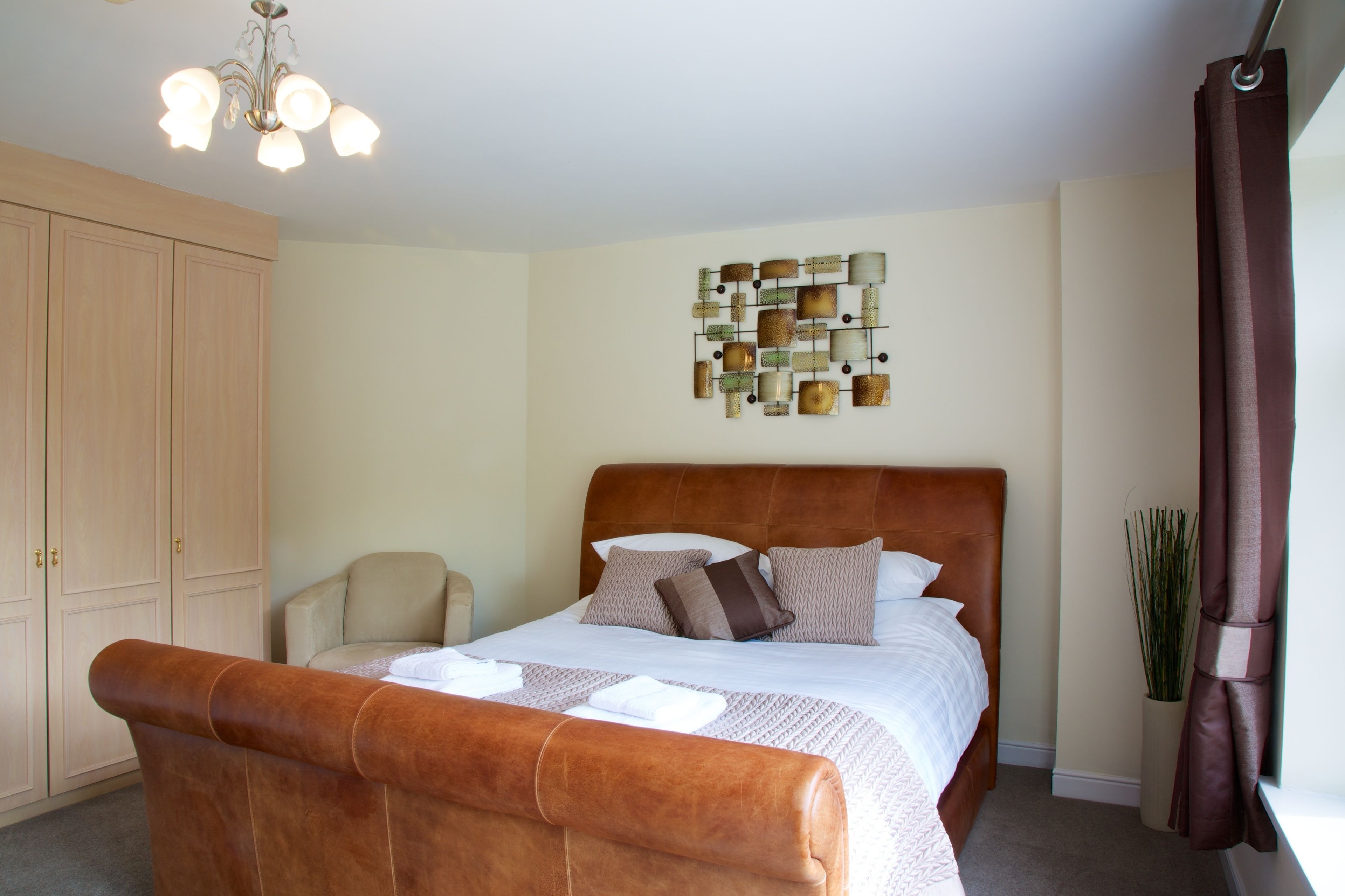 A bedroom with a large bed, a wooden wardrobe, a chair, a decorative wall piece, and a plant.