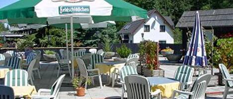 An outdoor seating area with white plastic chairs and tables, a green umbrella, and a house in the background.