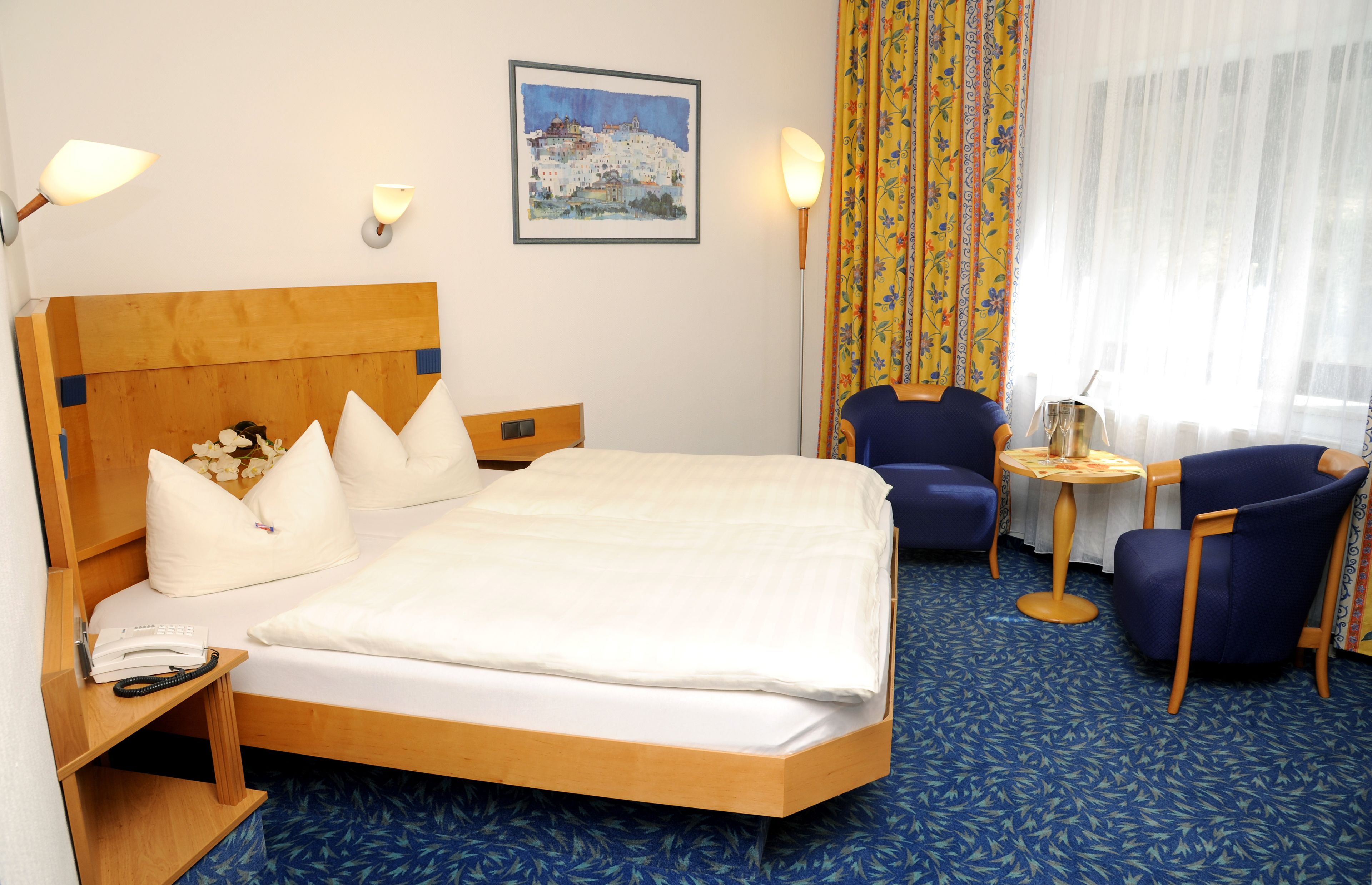 A hotel room with a large bed, a wooden headboard, a small table with a lamp, a chair, and a painting on the wall.