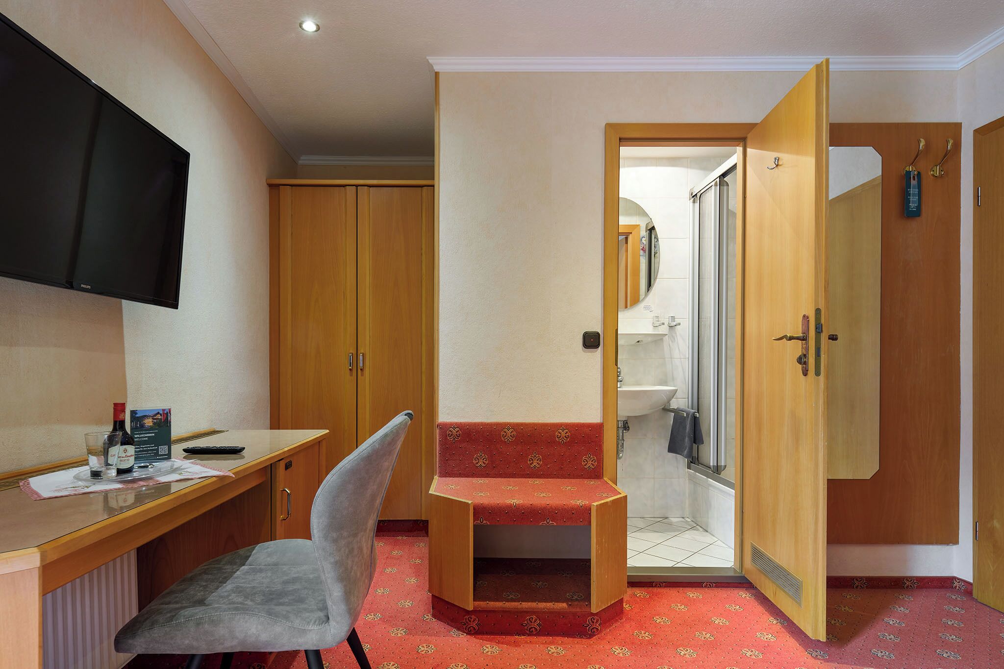 A hotel room with a wooden desk, a chair, a small red bench, a television, and a shower area.