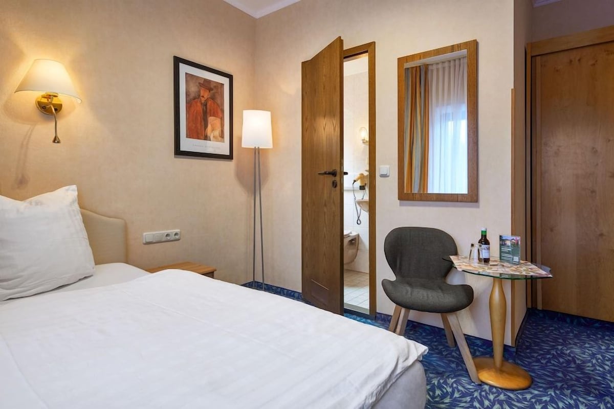 A hotel room with a bed, a chair, a table with a bottle and a menu, a framed picture on the wall, and a door leading to a bathroom.