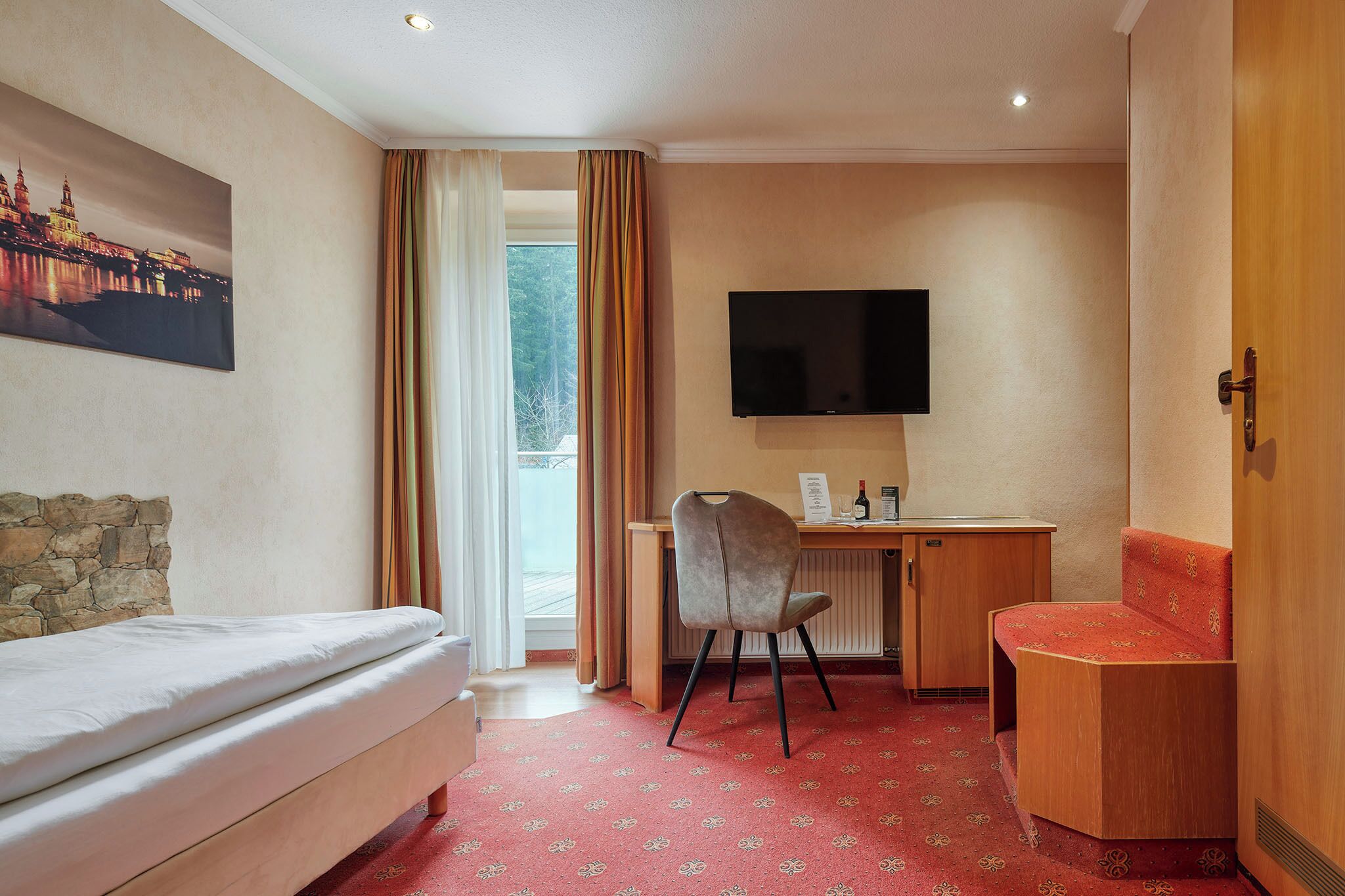 Habitación de hotel con cama, escritorio con silla, televisión y vista a la ciudad a través de una ventana.