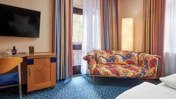 Habitación de hotel con televisor de pantalla plana, escritorio de madera con silla, sofá colorido y cama con ropa de cama blanca.