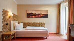 A hotel room with a bed, a nightstand, a lamp, a painting of a cityscape, and a window with curtains. - Hotel Ladenmühle (Altenberg)