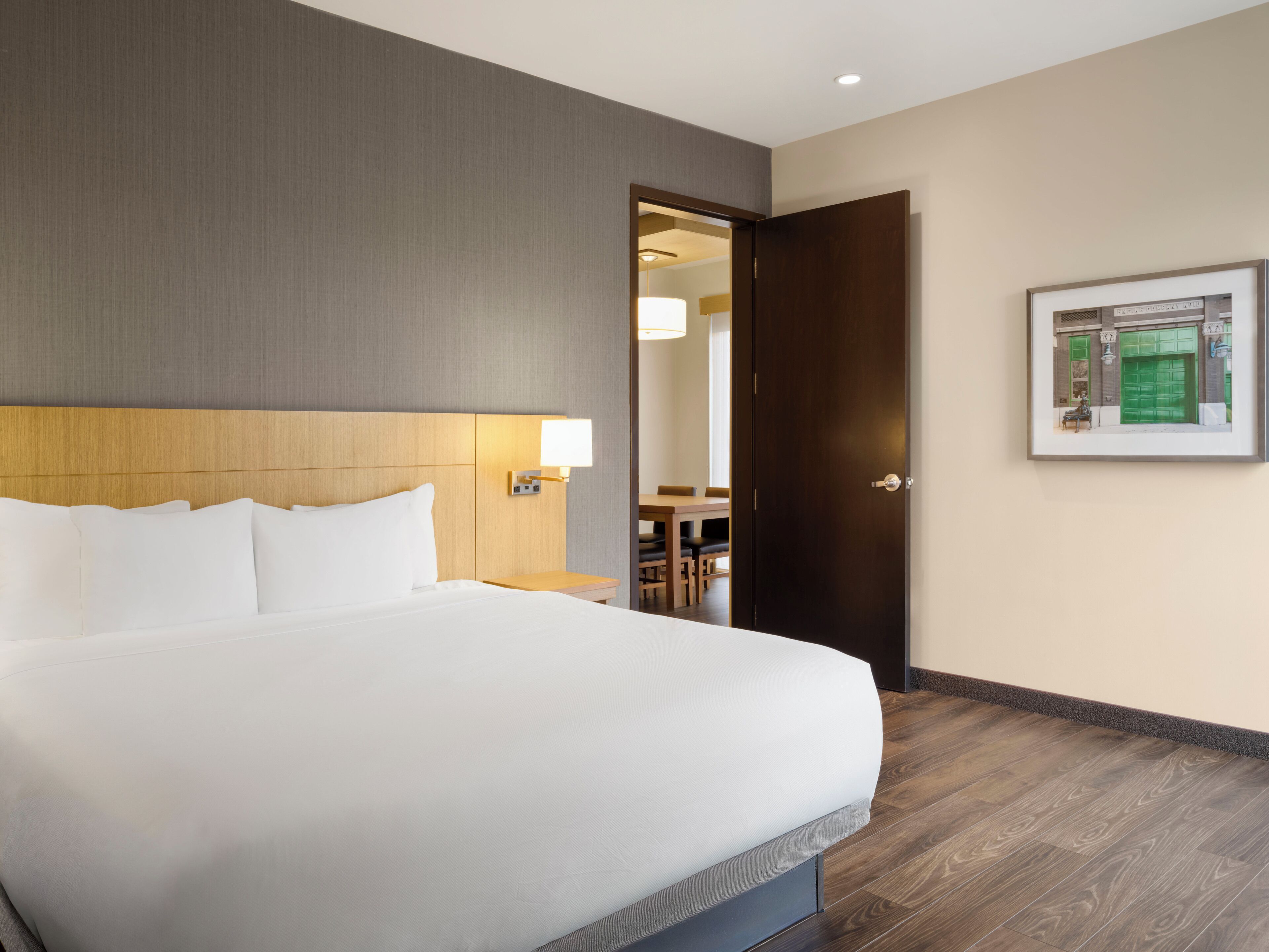 Hotelkamer met groot bed, houten hoofdbord, nachtkastje met lamp, deur naar een eetgedeelte, en een lijst aan de muur.