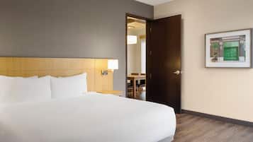 A hotel room with a large bed, a wooden headboard, a nightstand with a lamp, a door leading to a dining area, and a framed picture on the wall.