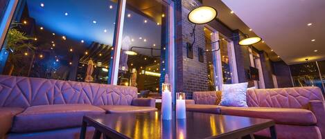 A modern outdoor seating area with cushioned sofas and a table, large windows, and ambient lighting.