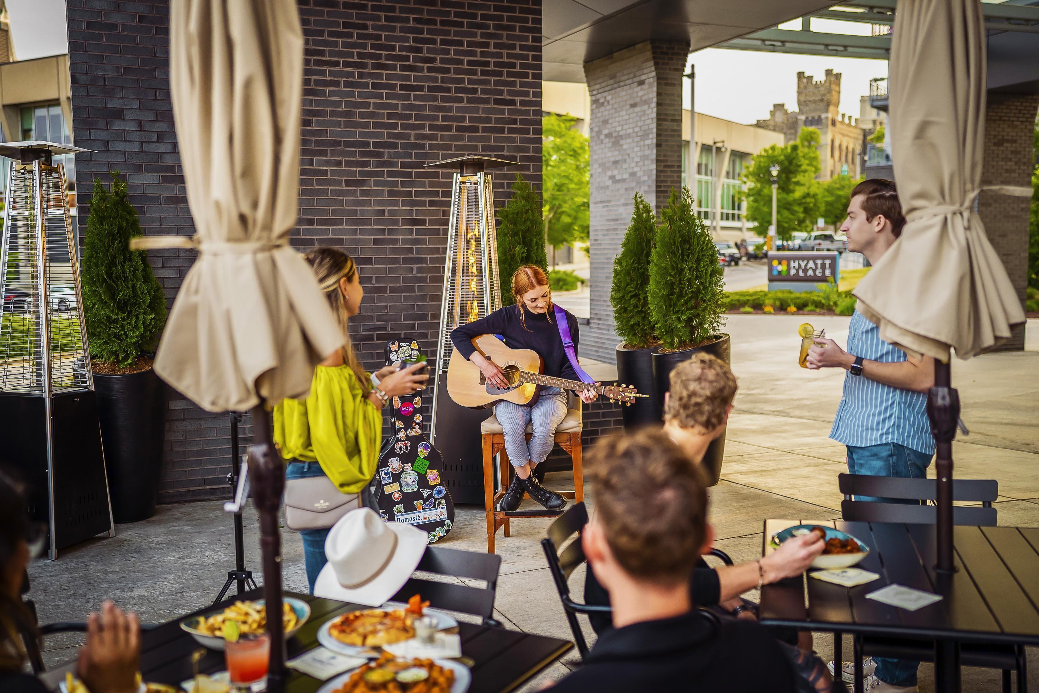 A musician performing at an outdoor seating area with tables and chairs, surrounded by potted plants and umbrellas.