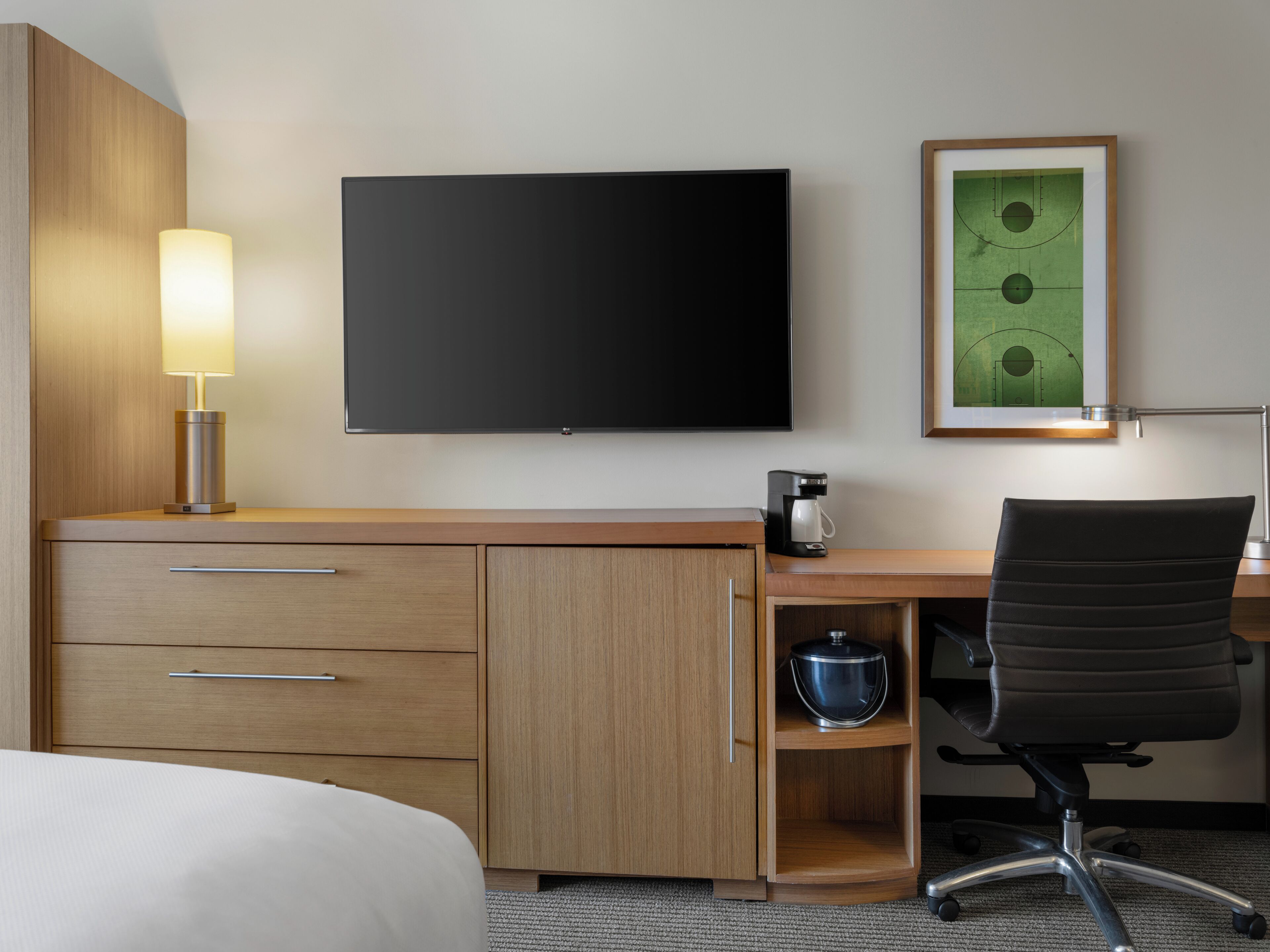A hotel room with a bed, a desk with a computer, a coffee maker, a chair, and a flat-screen TV mounted on the wall.