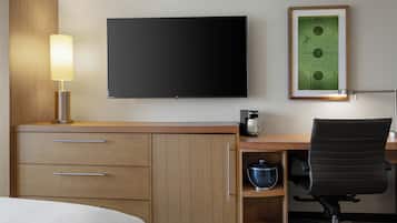 A hotel room with a bed, a desk with a computer, a coffee maker, a chair, and a flat-screen TV mounted on the wall.