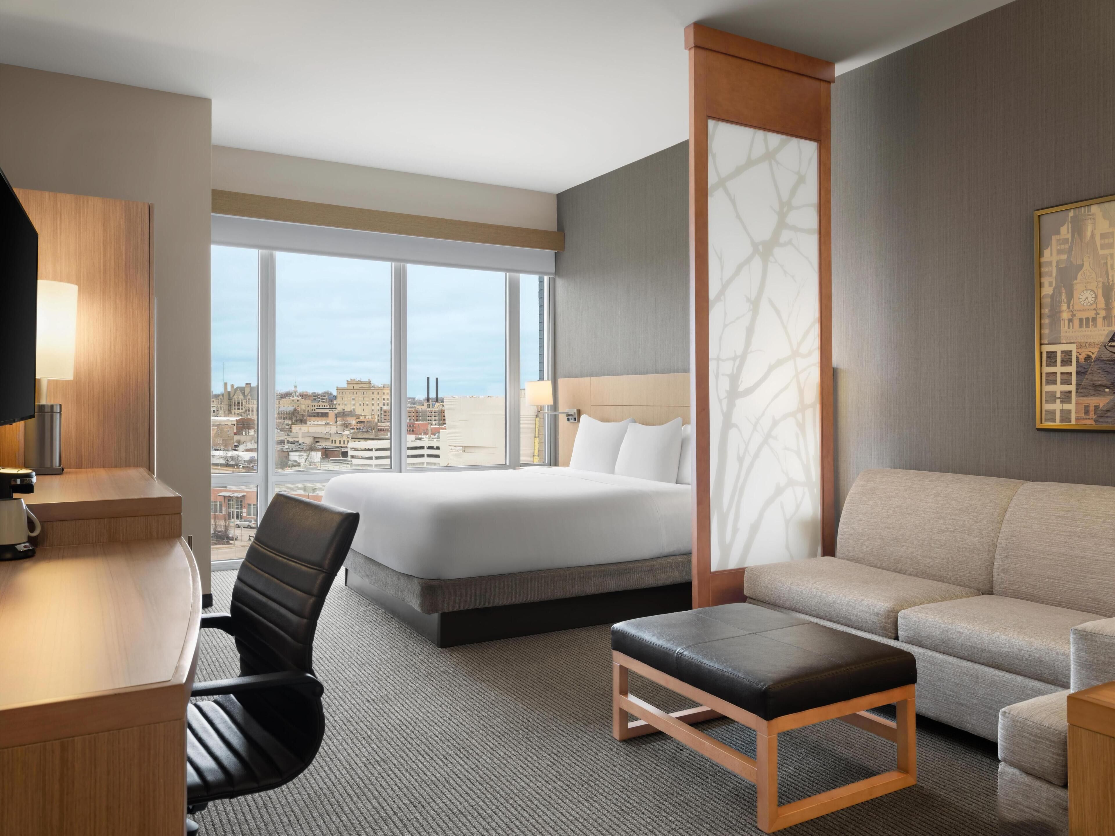 Une chambre d’hôtel comprenant un lit, un bureau avec une lampe, une chaise, un canapé et une grande fenêtre donnant sur la ville.