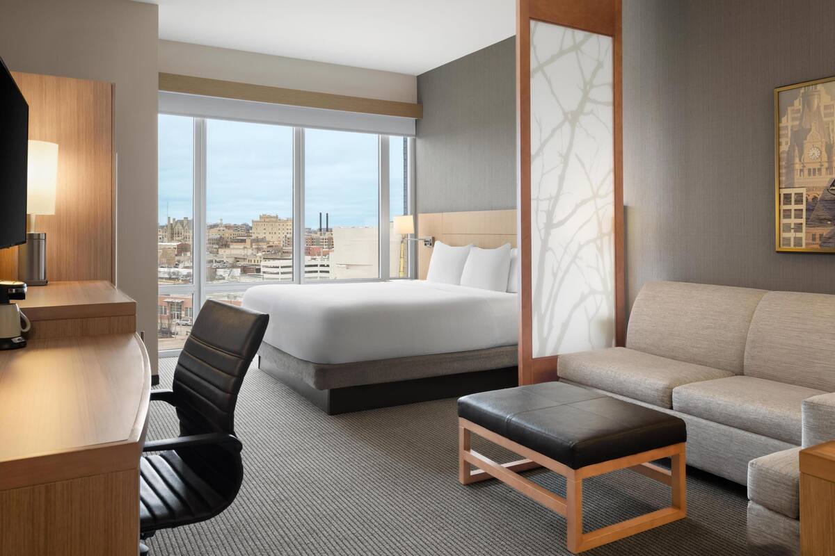A hotel room with a bed, a desk with a lamp, a chair, a sofa, and a large window offering a city view.