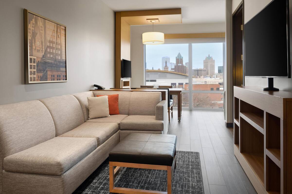 A modern living room with a sectional sofa, a coffee table, a TV, and a large window offering a city view.