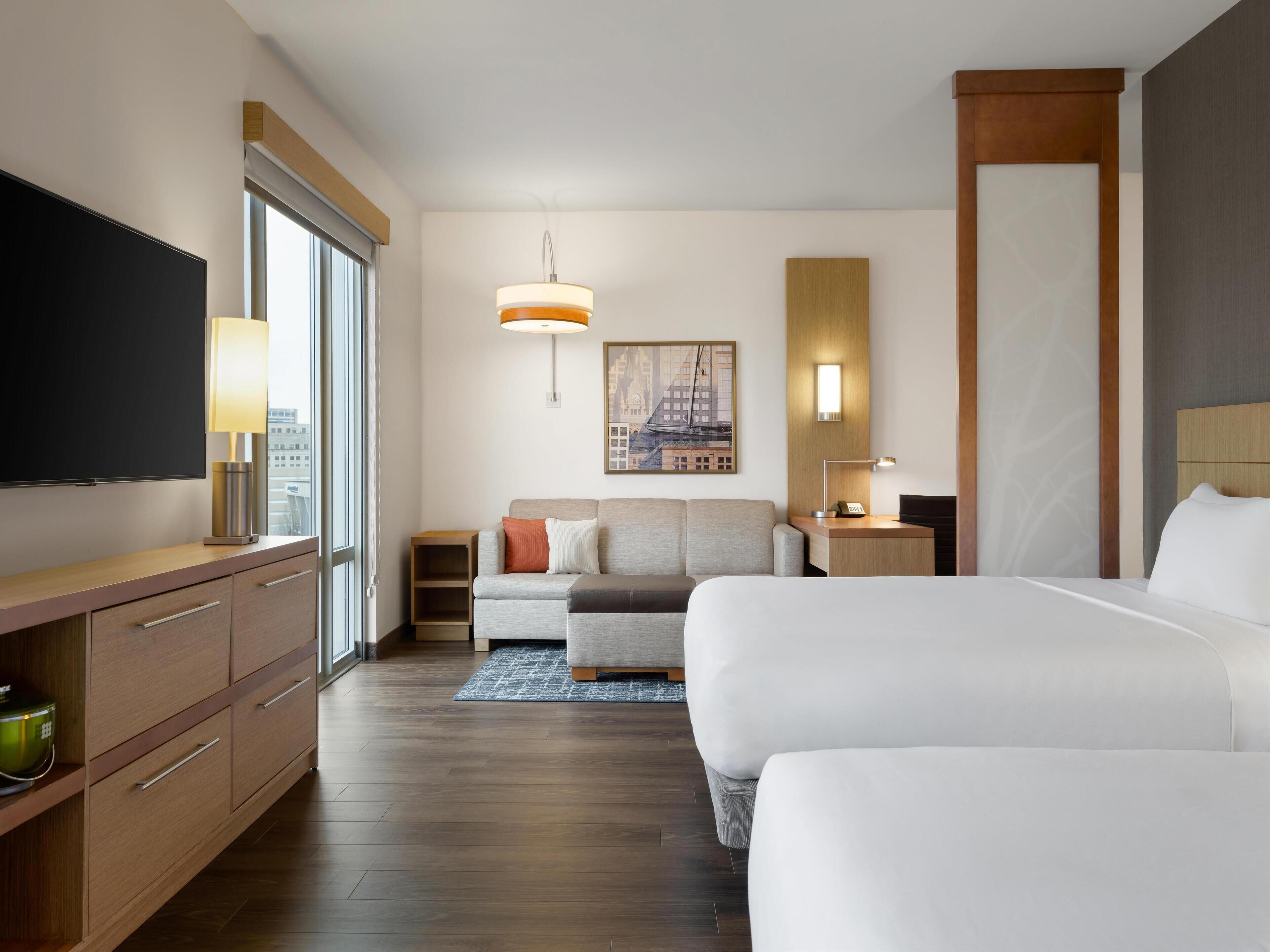 A hotel room with a large bed, a smaller sofa, a desk with a lamp, a TV on a cabinet, and a cityscape painting on the wall.