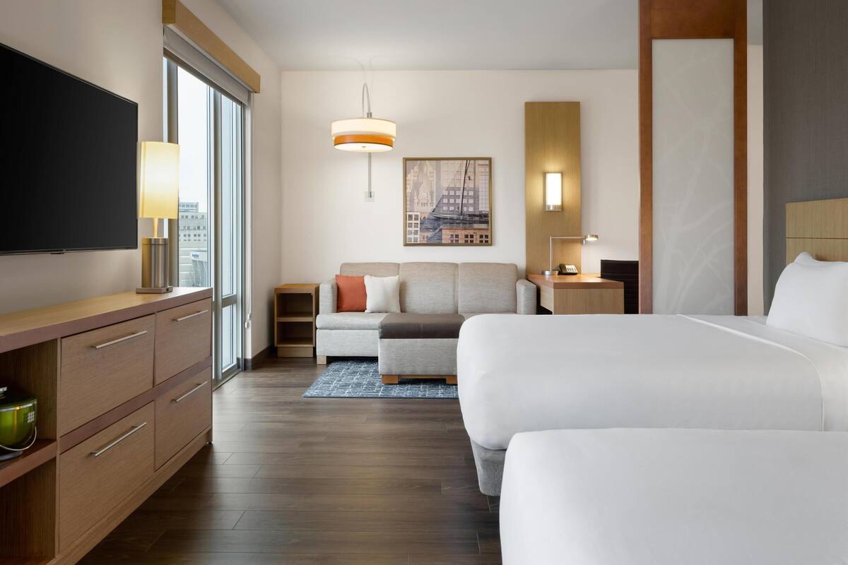 A hotel room with a large bed, a smaller sofa, a desk with a lamp, a TV on a cabinet, and a cityscape painting on the wall.