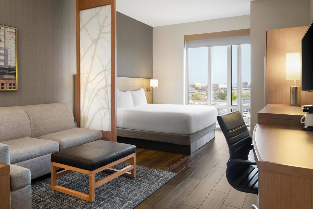A hotel room with a bed, a sofa, a desk, and a view of the city through large windows.