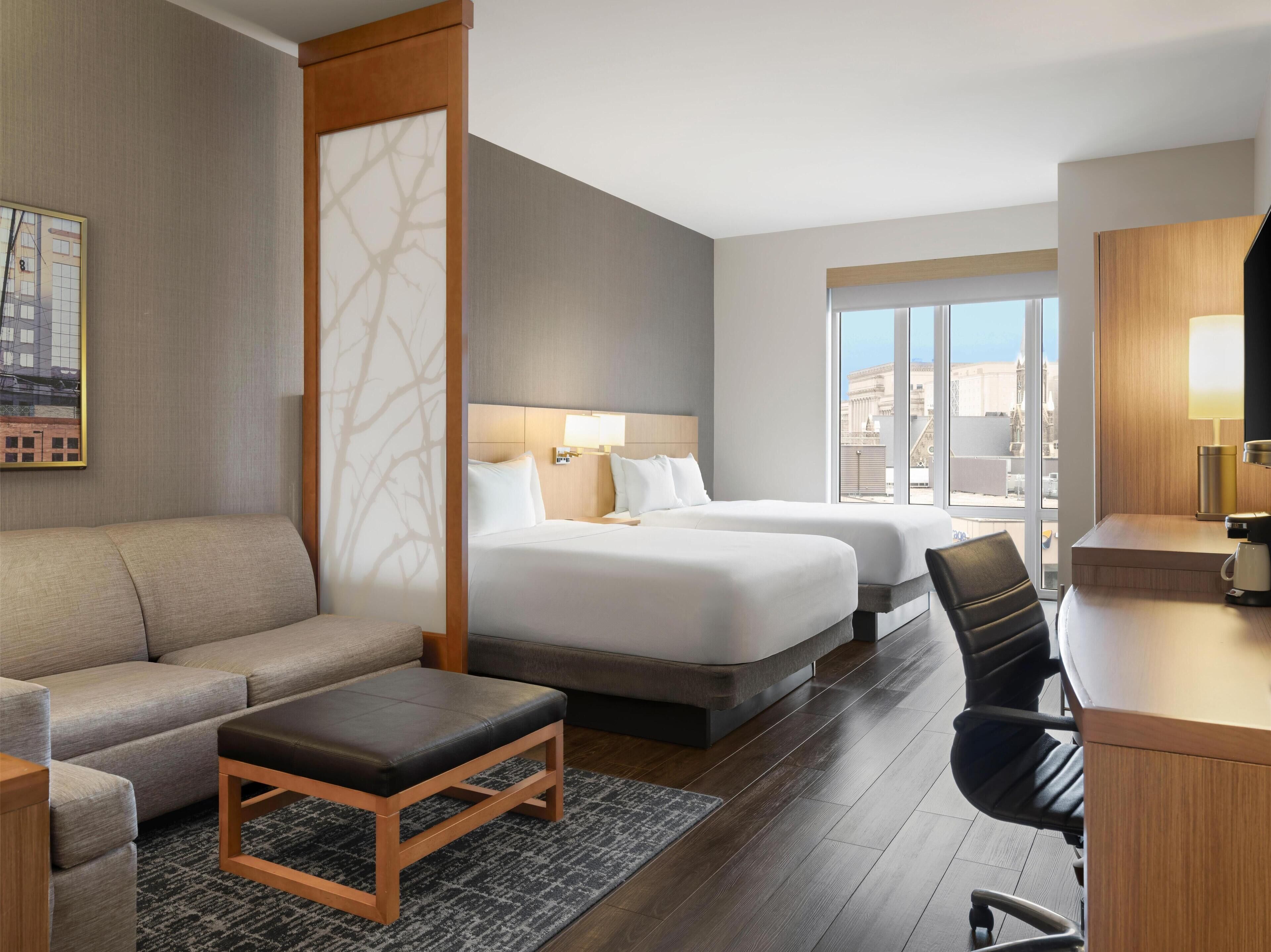 A hotel room with a sofa, two beds, a desk, and a view of the city.