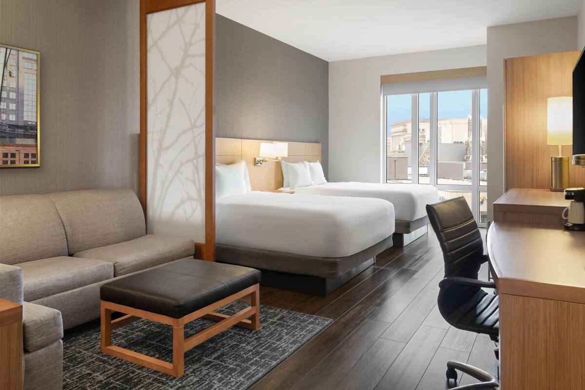 A hotel room with a sofa, two beds, a desk, and a view of the city.