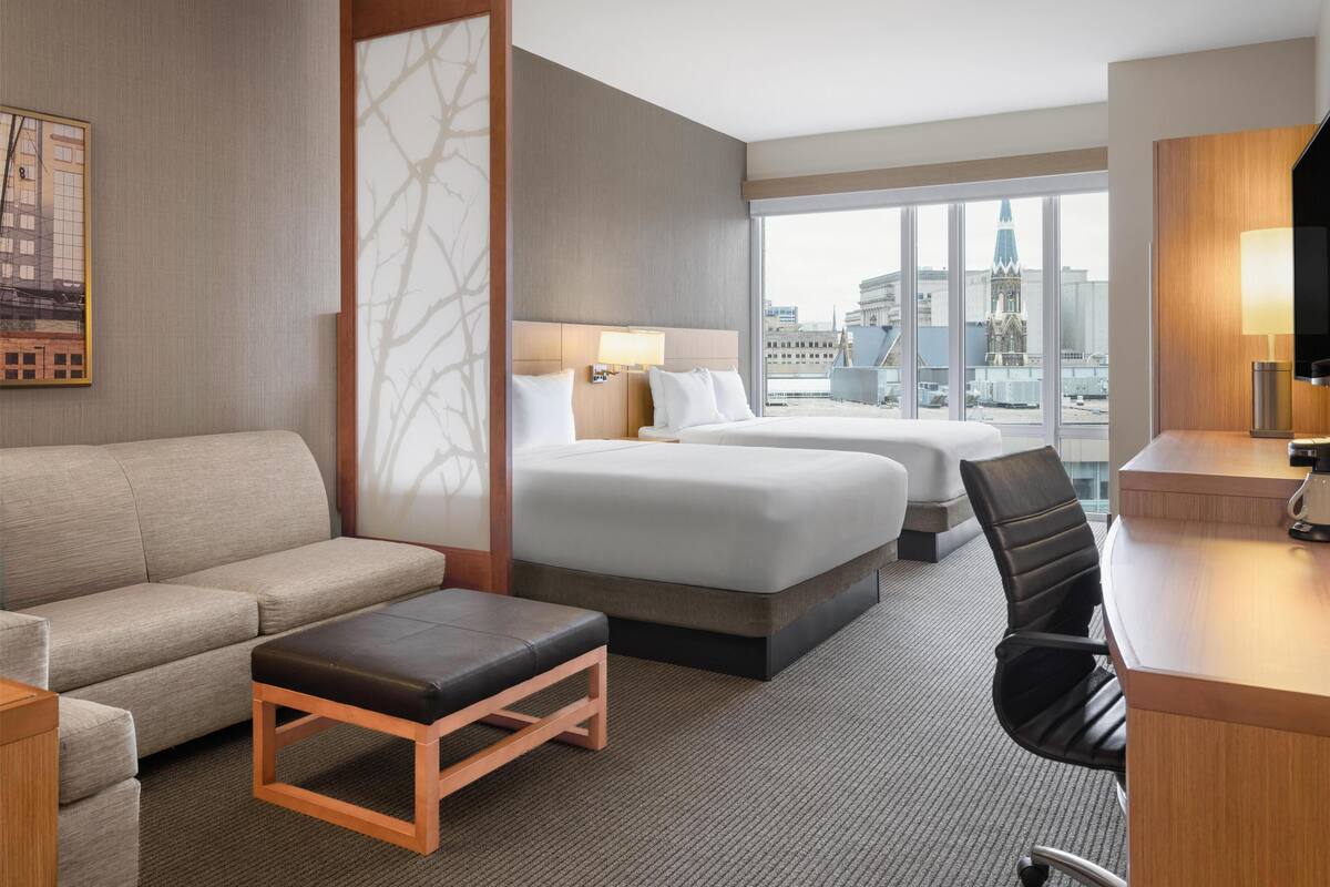 A hotel room with a sofa, two beds, a desk, and a view of the cityscape.