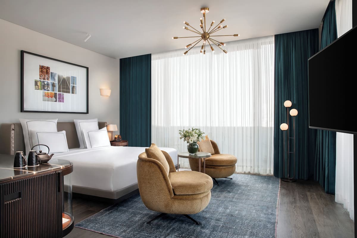 A modern hotel room with a large bed, two armchairs, a desk, and a television.