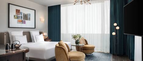 A modern hotel room with a large bed, two armchairs, a desk, and a television.