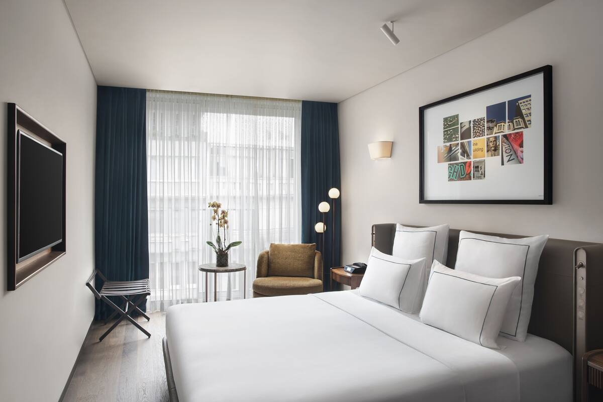 A hotel room with a large bed, a chair, a small table with a plant, a bedside table with a lamp, and a framed collage on the wall.
