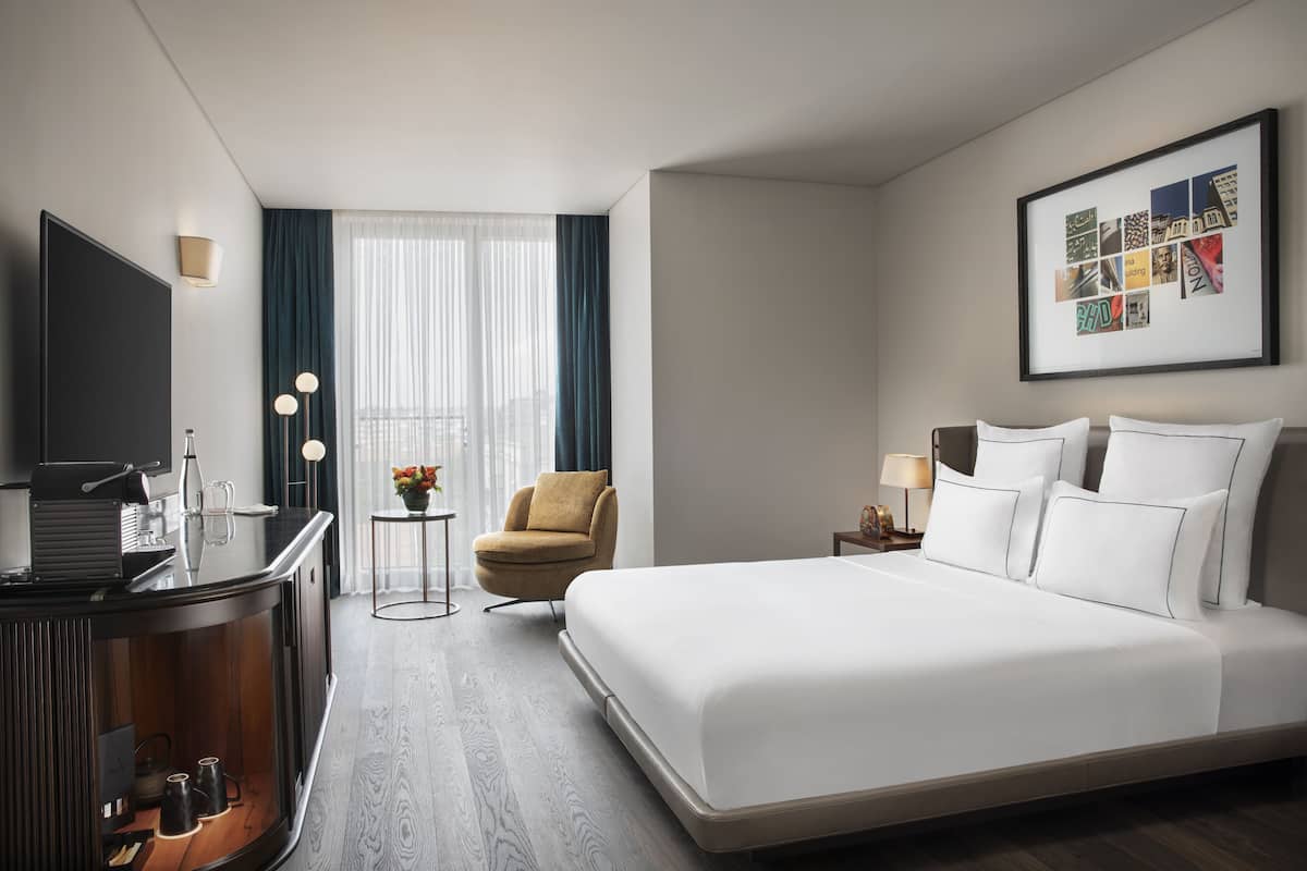 A hotel room with a large bed, a desk with a flat-screen TV, a chair, a small table with a lamp, and a framed collage on the wall.