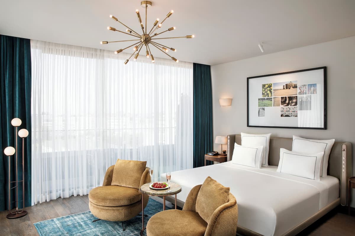 A hotel room with a large bed, two armchairs, a round table with a plate of fruit, a chandelier, a framed picture on the wall, and a floor lamp.