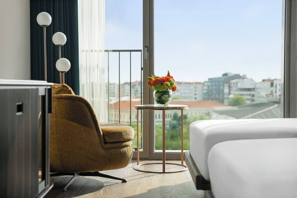 A hotel room with a bed, a chair, a small table with a vase of flowers, and a view of buildings through a large window.