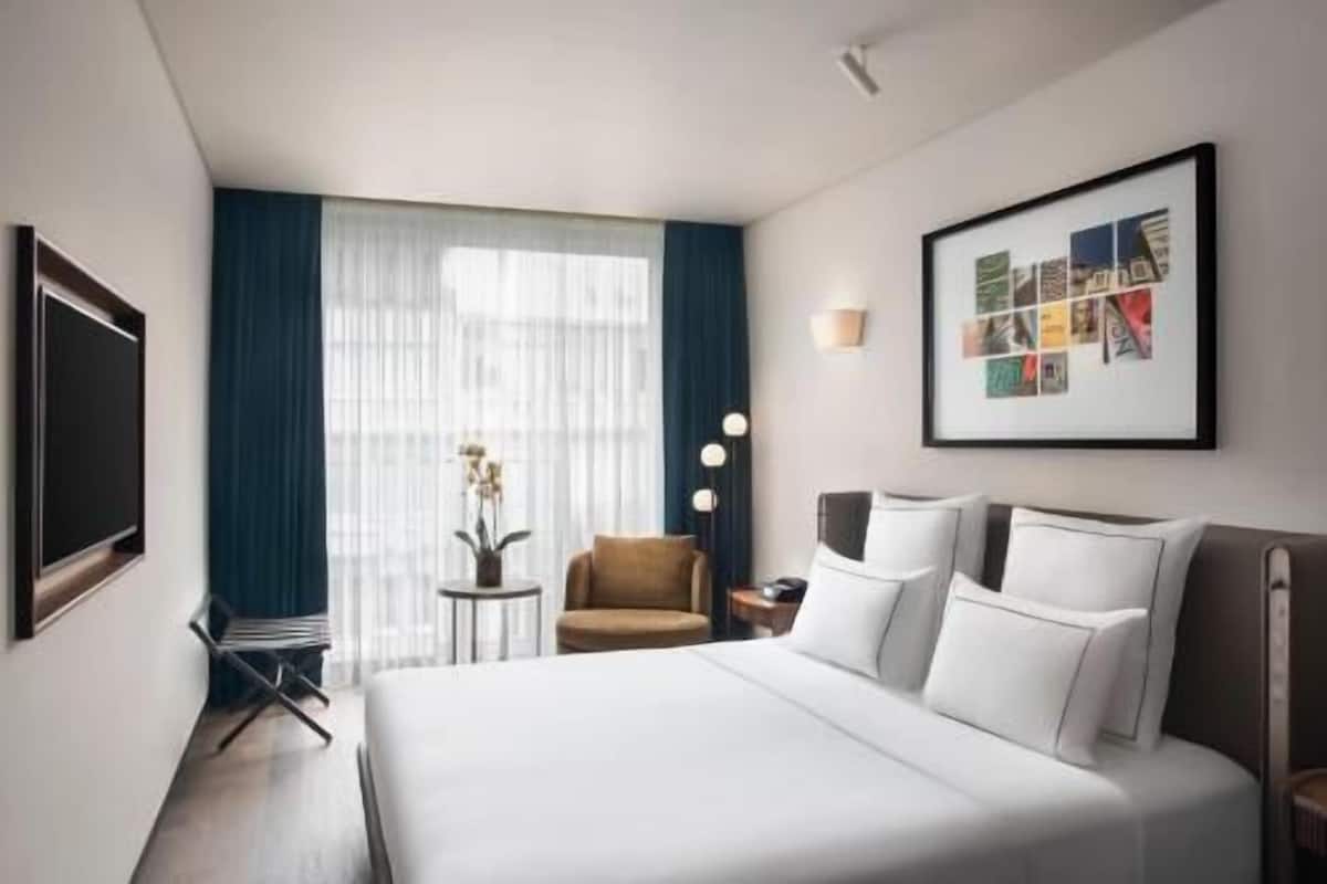 A hotel room with a bed, a chair, a small table with a vase, a wall-mounted TV, and a framed picture.