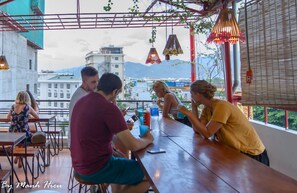Un grupo de personas sentadas en una mesa de madera en un espacio de comedor al aire libre con vistas a la ciudad, bajo lámparas colgantes.