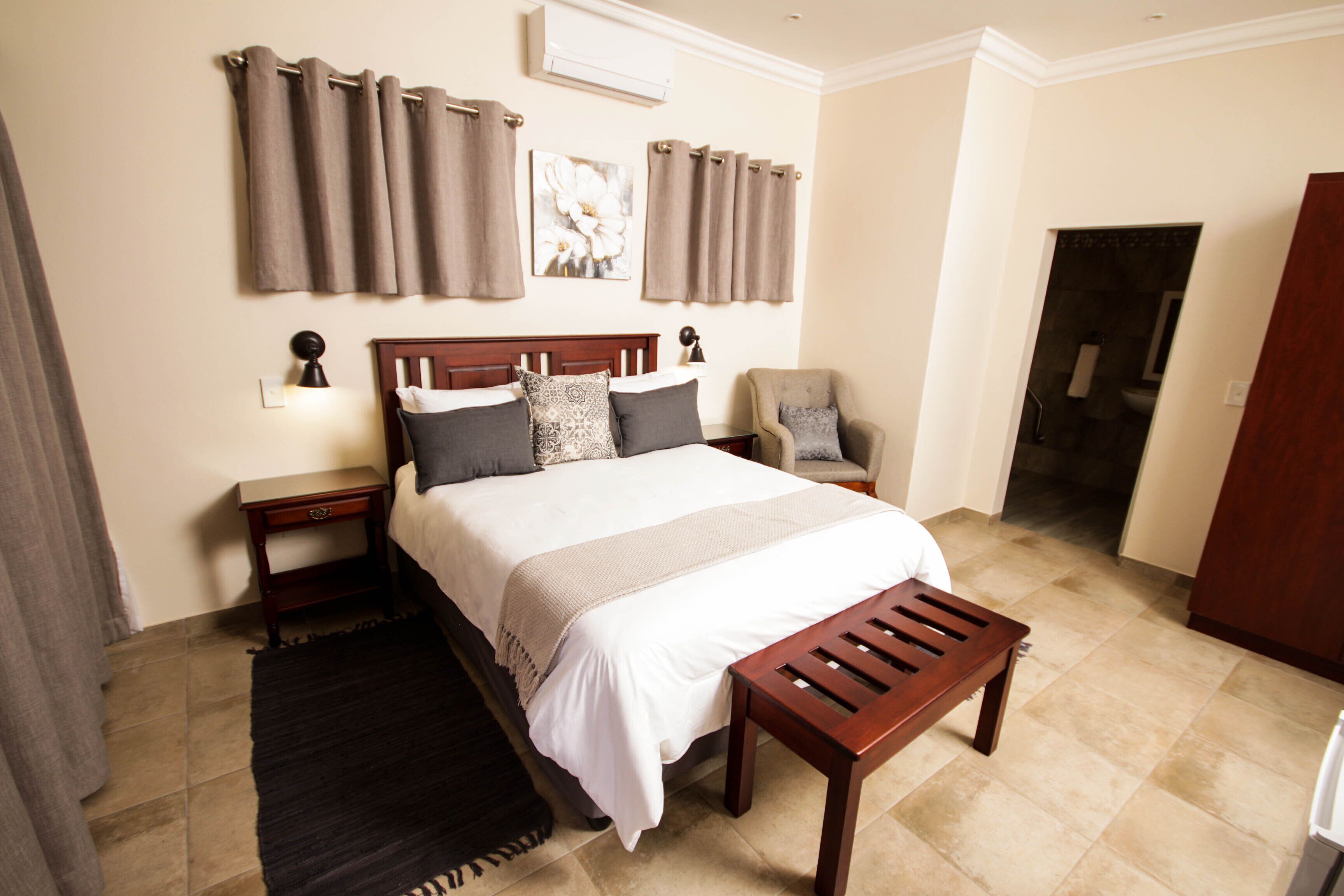 A bedroom with a bed, bedside tables, a chair, a wooden bench, curtains, and a painting on the wall.