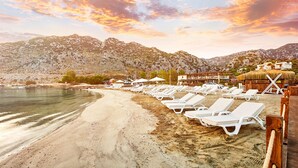 A beach with sun loungers and umbrellas, a pier, and mountains in the background.