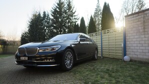 Un sedán BMW negro estacionado en una entrada con un muro de ladrillo y una cerca de madera al fondo.