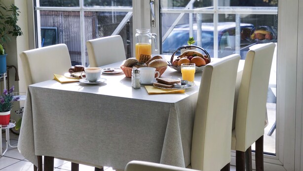 Una mesa de desayuno con diversos alimentos, una jarra de jugo de naranja y vista a coches estacionados y follaje en el exterior.