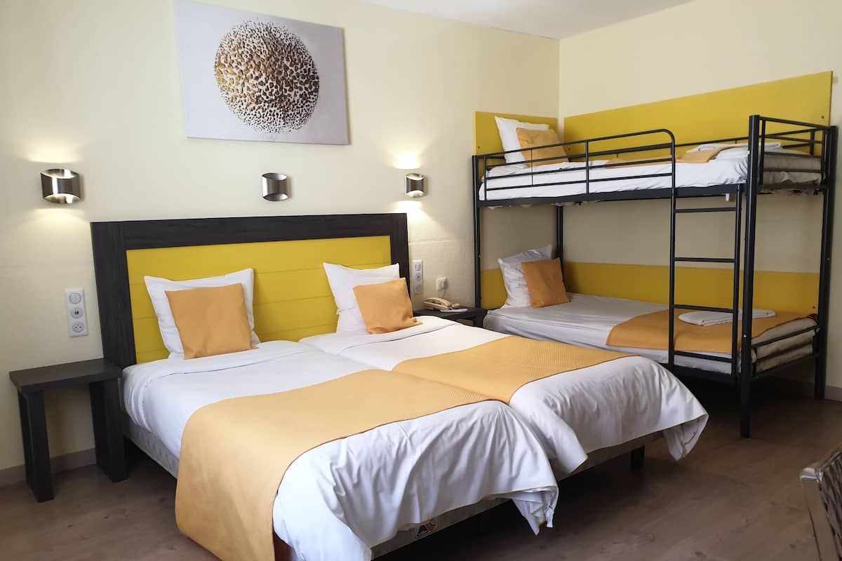 Une chambre d’hôtel avec deux lits simples et un lit superposé, tous dotés de têtes de lit jaunes. On y trouve une table de chevet avec une lampe, une prise de courant et un téléphone fixé au mur.