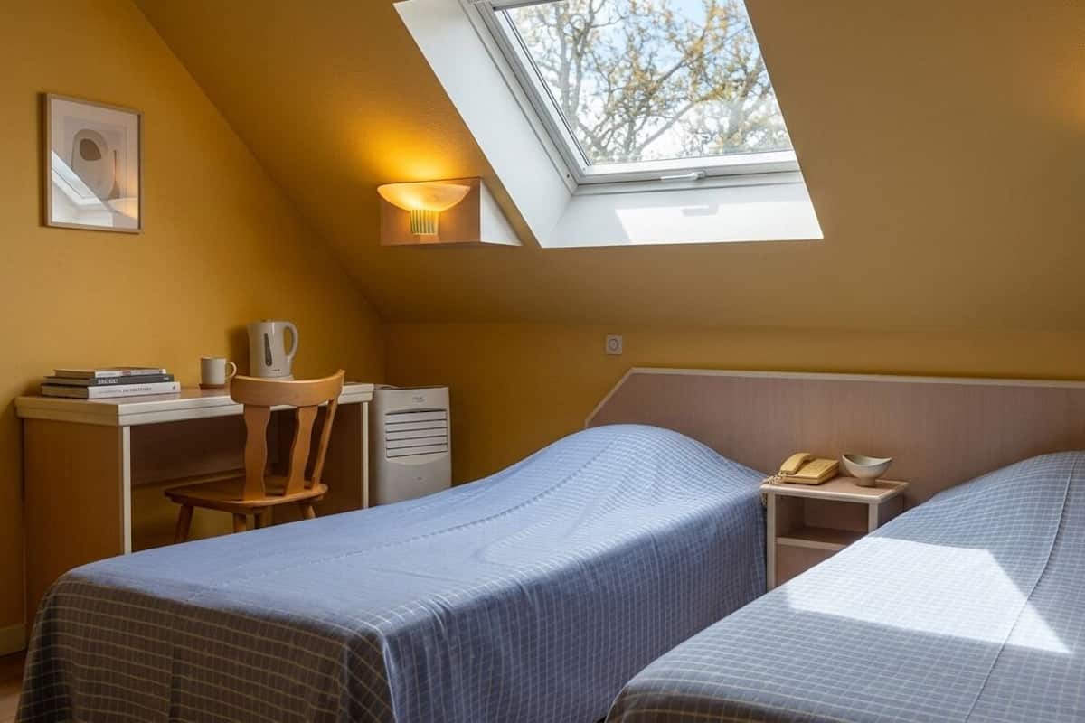 Une chambre avec deux lits, un bureau, une chaise, une lucarne et une table de chevet avec une lampe.