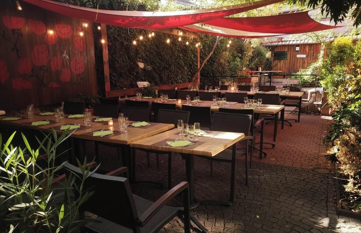 Un área de comedor al aire libre con mesas preparadas para dos, rodeada de follaje y un muro de madera con un mural floral.