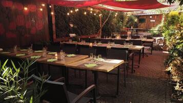 Un área de comedor al aire libre con mesas preparadas para dos, rodeada de follaje y un muro de madera con un mural floral.
