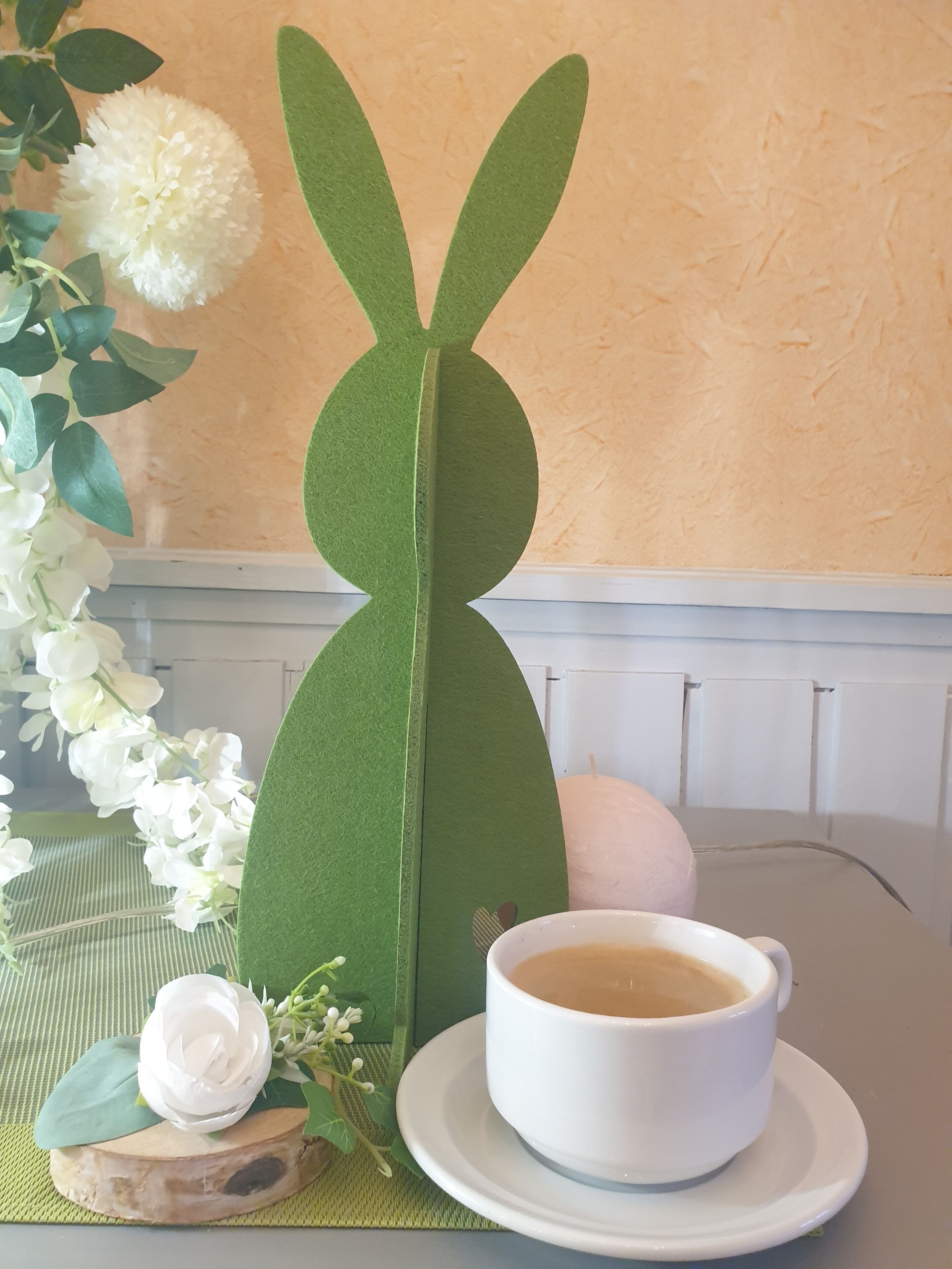Una mesa con una taza de café blanco, un adorno de conejo verde y flores blancas.