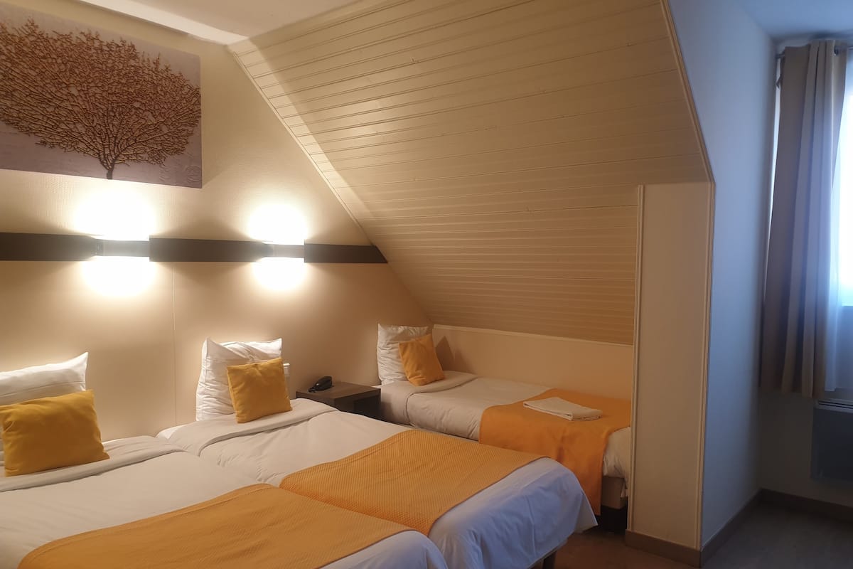 Une chambre d’hôtel avec deux lits, chacun agrémenté de draps blancs et de coussins décoratifs orange. Un luminaire est fixé au mur au-dessus des lits, et un tableau représentant un arbre est accroché au mur.
