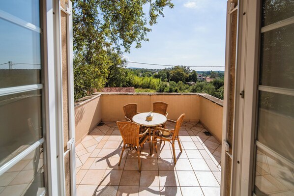 Un balcon avec une table ronde et des chaises en osier, donnant sur un jardin et des bâtiments situés au loin.