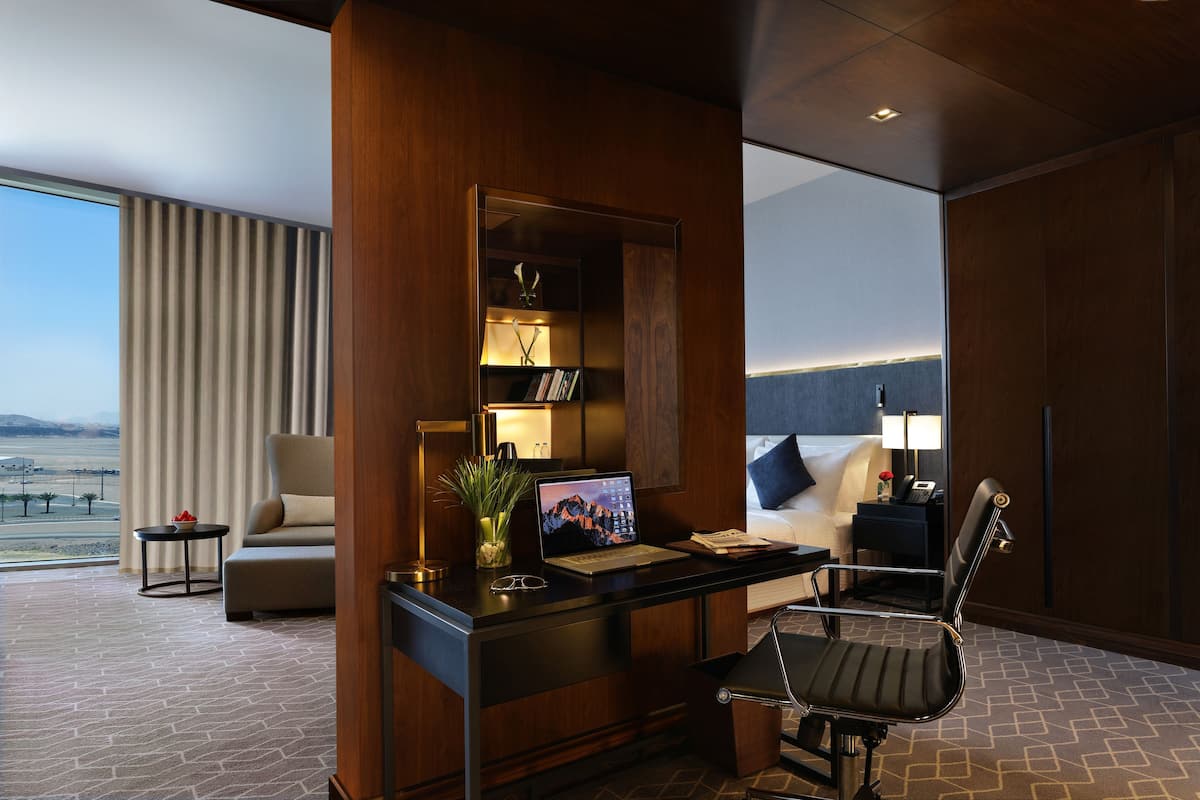 Ein modernes Hotelzimmer mit großem Fenster, einem Schreibtisch mit Laptop, einer gemütlichen Couch, einem Sessel und einem Nachttisch mit Lampe.