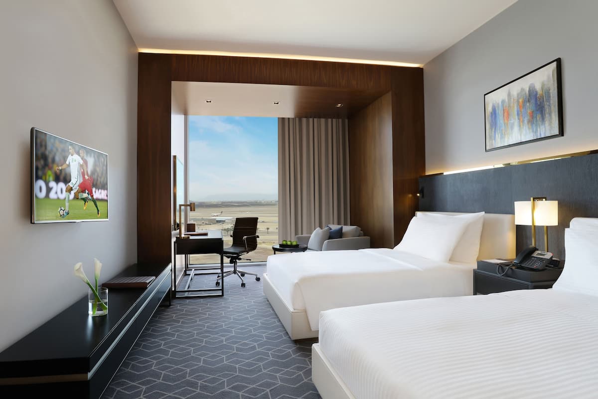 Ein Hotelzimmer mit zwei Betten, einem Schreibtisch, einem Sessel, einem Fernseher und Blick ins Freie.